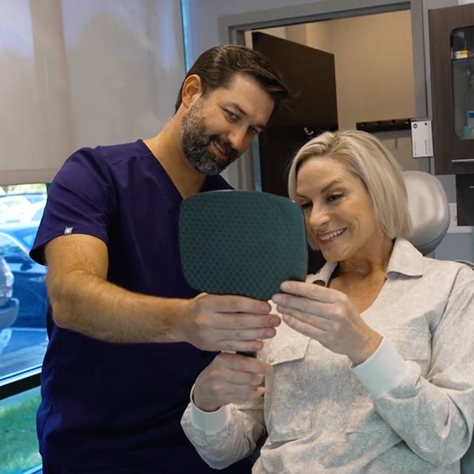 Dr. Morelli showing a Raleigh patient her new smile in a mirror after a successful All-on-4 dental implant procedure.