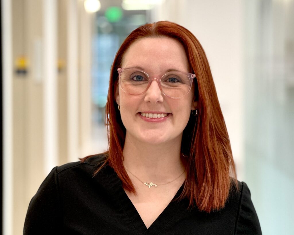 A head-and-shoulders portrait of Callie Howlett, a smiling Dental Assistant with bright red hair and pink glasses, wearing black FIGS scrubs in a modern dental office hallway.