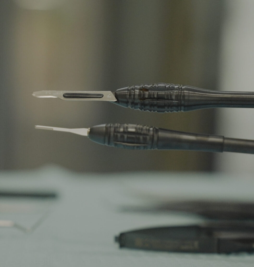 A close-up photograph comparing a larger traditional scalpel (top) with a smaller, more refined micro-scalpel (bottom), both featuring black ergonomic handles and lying horizontally over other blurred dental tools on a sterile blue surface.