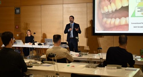 Dr. Thiago Morelli standing on stage and speaking into a microphone during a professional lecture. He is wearing a dark suit and a conference lanyard, gesturing with his hand as he presents. Behind him, a large projection screen displays a clinical dental photo of a patient's teeth and a diagram illustrating periodontal surgical techniques like MIST and M-MIST. In the foreground, several conference attendees are seated at long workshop tables listening to the presentation inside a wood-paneled lecture hall.