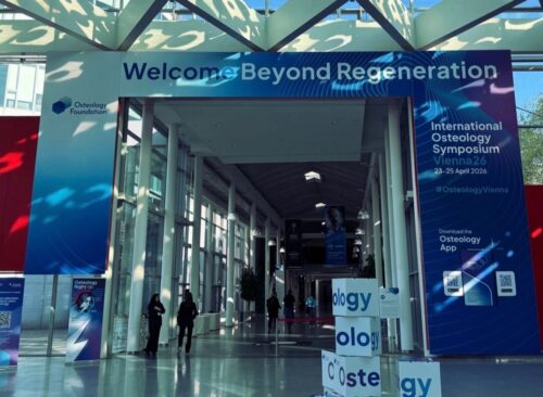 Wide-angle photograph of the brightly lit main entrance to the International Osteology Symposium in Vienna. Large blue and red banners flank the entrance, displaying 'Welcome Beyond Regeneration', 'International Osteology Symposium Vienna26', and the 'Osteology Foundation' logo. Stacked cubes in the foreground spell out 'Osteology'. Conference attendees are visible in the background under high skylights.
